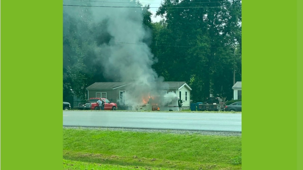 A house with a fire and thick gray smoke rising from the roof, with a red pickup truck and trees in the background.