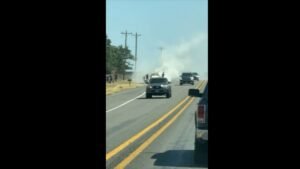 A black SUV on a two-lane road with smoke rising in the distance, viewed from another vehicle.