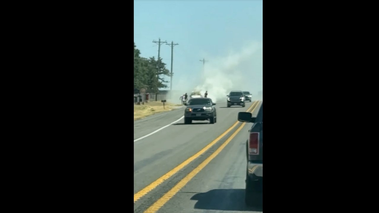 A black SUV on a two-lane road with smoke rising in the distance, viewed from another vehicle.