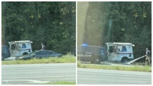 A damaged white delivery van and a black police SUV are stopped beside a highway, with a person spraying water near the van and trees in the background.