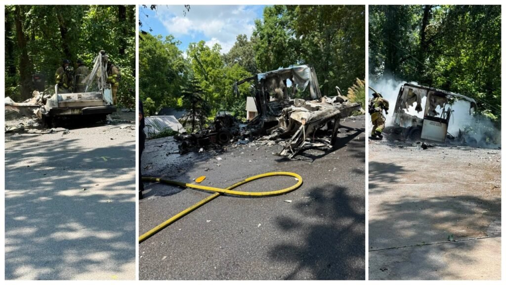 A burned-out vehicle sits on a tree-lined road, with debris scattered across the pavement and a yellow hose stretched in front.