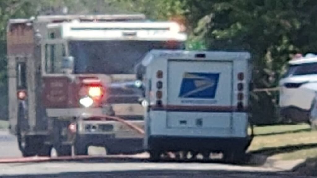 An emergency vehicle with flashing lights is parked behind a white USPS mail truck on a residential street.