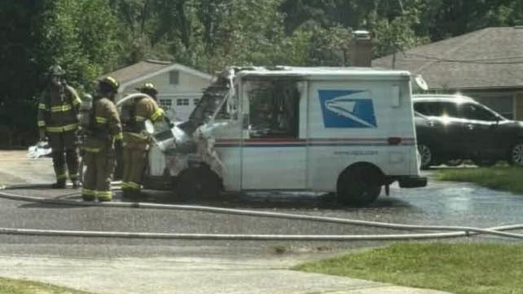 A damaged mail truck with a burned front end sits on a wet residential street as firefighters stand nearby, with houses and trees in the background.