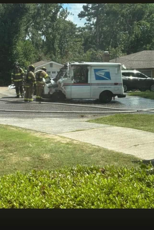Firefighters work beside a damaged USPS mail truck on a suburban street, with hoses on the pavement and houses and trees in the background.