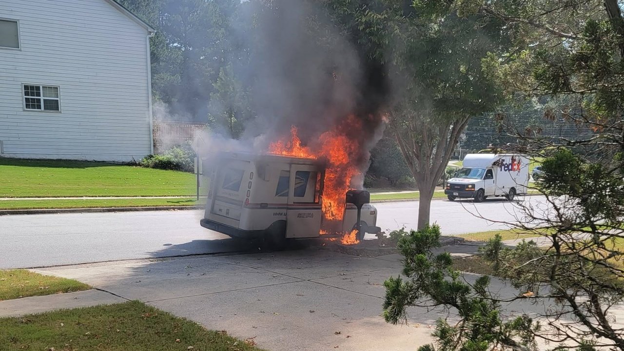 A FedEx mail truck burns on a residential street, with flames and thick smoke rising beside a driveway and a white house.