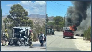 A burned mail truck sits on a roadside as firefighters stand nearby, with smoke rising behind a red car on the street.