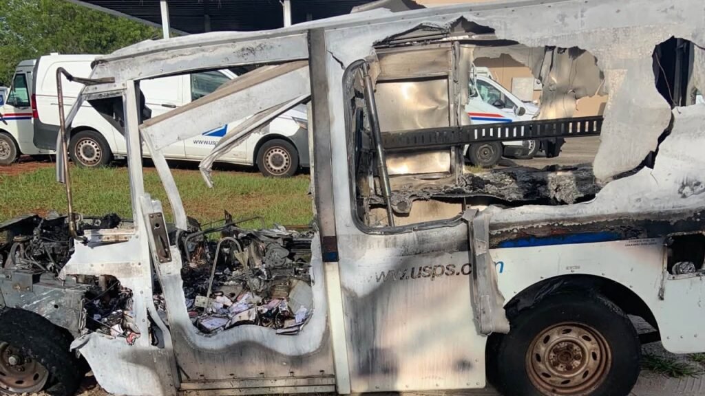 A burned-out white utility truck with its side panel and windows badly damaged, parked on grass with other vehicles in the background.