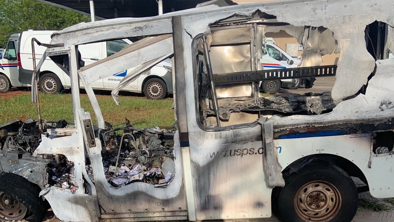 A burned-out white utility truck with its side panel and windows badly damaged, parked on grass with other vehicles in the background.