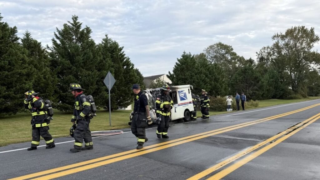 Firefighters stand beside a USPS mail truck on the shoulder of a rural road, with a hose stretched across the wet pavement and trees lining the background.