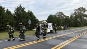 Firefighters stand beside a USPS mail truck on the shoulder of a rural road, with a hose stretched across the wet pavement and trees lining the background.