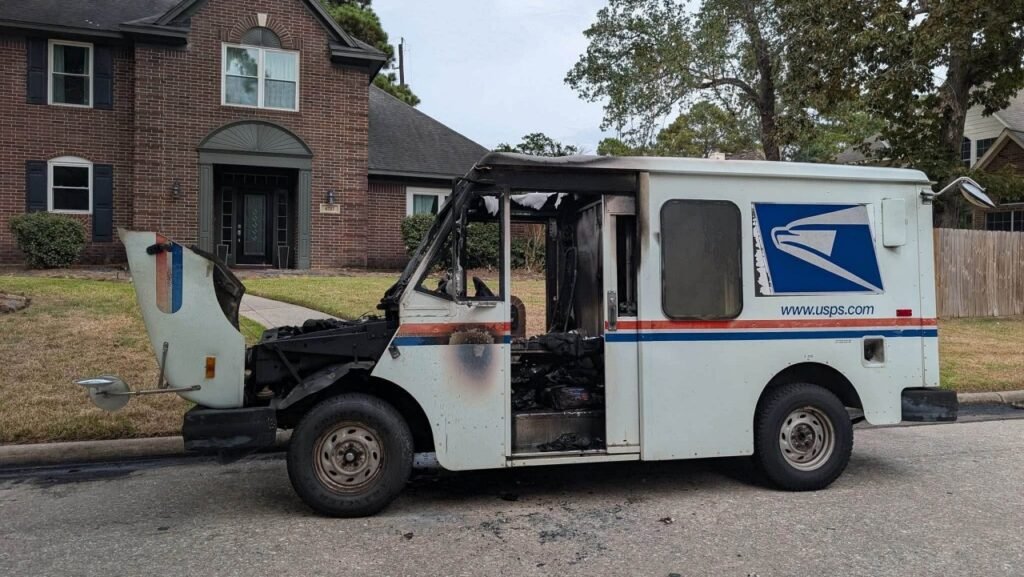 A burned USPS mail truck is parked on a residential street, with the front cab and side door heavily damaged in front of a brick house.