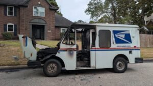 A burned USPS mail truck is parked on a residential street, with the front cab and side door heavily damaged in front of a brick house.