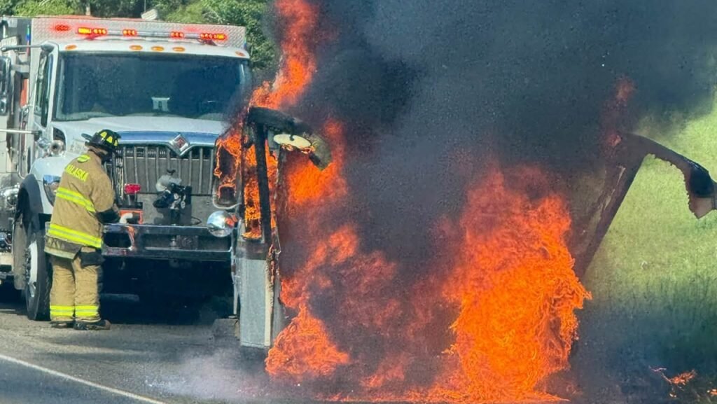 A mail truck is engulfed in flames and thick black smoke on a roadside while a firefighter stands nearby in front of an emergency vehicle.