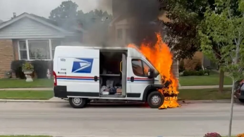 A USPS mail truck burns on a suburban street, with flames coming from the front passenger side and smoke rising above the vehicle.