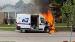 A USPS mail truck burns on a suburban street, with flames coming from the front passenger side and smoke rising above the vehicle.