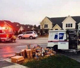 A US postal service truck damaged and partially burnt roadside with packages scattered nearby. A fire truck is parked in response to the incident, with emergency lights illuminated, while residential houses are visible in the background.