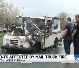 7Av CPU77W0 1 Two individuals observing a damaged mail truck that was affected by a fire, with noticeable burn marks and smoke damage. The scene is set in a parking area with trees nearby.