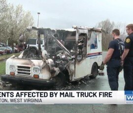 7Av CPU77W0 1 Two individuals observing a damaged mail truck that was affected by a fire, with noticeable burn marks and smoke damage. The scene is set in a parking area with trees nearby.