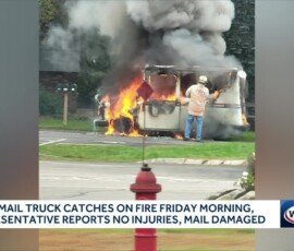 A USPS mail truck on fire with flames and smoke rising, parked in a lot. Nearby, a person wearing a white shirt is standing. A fire hydrant is visible in the foreground. No injuries reported.