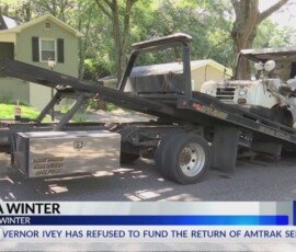 A tow truck with a flatbed is positioned on a residential street, transporting a small bus. A house is visible in the background, along with trees providing shade.
