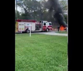 A fire truck is parked in a grassy area while thick black smoke rises from a nearby location, indicating a fire. Trees are visible in the background.