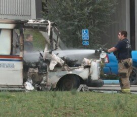 A firefighter using a hose to extinguish flames on a damaged United States Postal Service vehicle. Smoke and charred surfaces are visible. A blue pickup truck is parked in the background.