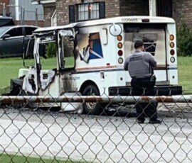 A damaged postal service vehicle with burn marks parked on a street. A police officer is standing nearby while a person in a white outfit observes from a distance. A residential area is visible in the background.