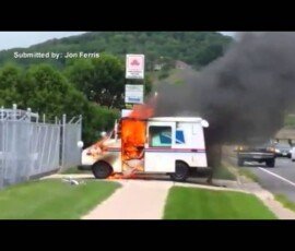 A postal delivery vehicle is on fire, with flames and thick black smoke coming from the rear. The scene includes a sidewalk and a grassy area, along with a sign in the background.