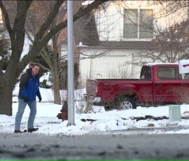 A man wearing a blue jacket walking on a snowy street, with a red pickup truck parked nearby. Trees and houses are visible in the background, indicating a residential neighborhood.
