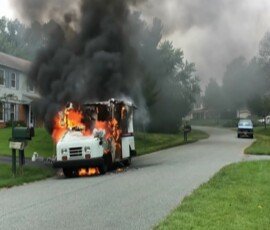 A vehicle engulfed in flames on a residential street, with dense black smoke billowing into the air. Houses and a parked car are visible in the background.