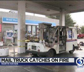 A damaged mail truck at a gas station showing signs of a fire, with charred areas on the vehicle. In the background, a Mobil Mart sign is visible along with several vehicles parked nearby.