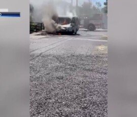 A vehicle partially engulfed in flames, emitting smoke, in a gravel area near a gate. Firefighters can be seen responding in the background.