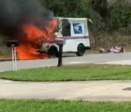 A USPS delivery vehicle engulfed in flames while emitting thick black smoke, parked near a mailbox on a residential street. In the background, grassy areas and trees are visible.