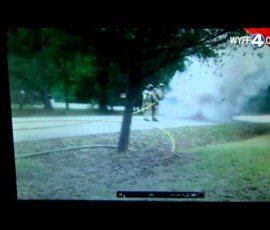 Firefighters responding to an incident near a residential area, with smoke rising from the ground. A fire hose is laid out on the grass, and trees are visible in the background.