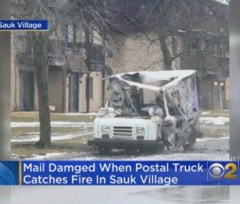 A damaged postal truck with a charred front, parked on a wet street in Sauk Village. Nearby structures are visible in the background, with overcast weather conditions.