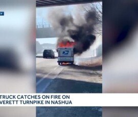 A USPS truck engulfed in flames on the side of the Everett Turnpike in Nashua, with thick black smoke rising into the air. An overpass is visible in the background.